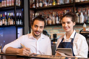 Happy young waiter making notes in notepad with his pretty colleague near by