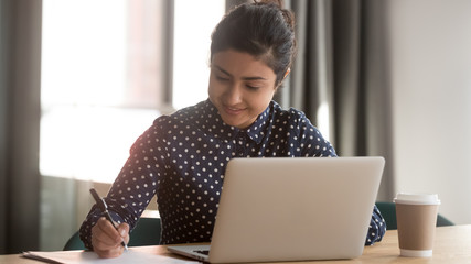 Indian businesswoman hold ballpoint signing documents sitting at desk in workplace using computer business application, concentrated attractive employee make notes prepare report busy workday concept