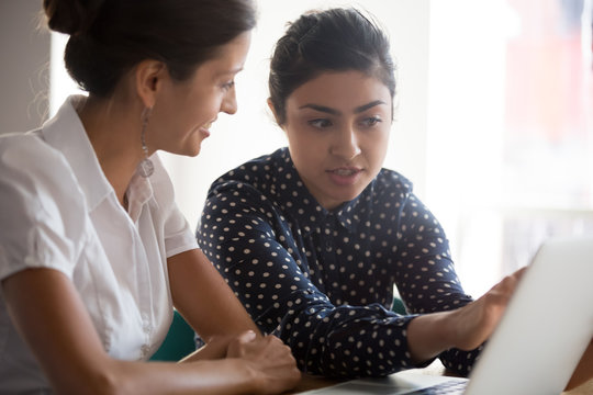 Diverse businesswomen discuss analyse online project look at laptop screen colleagues brainstorming strategizing together, mentor helping apprentice explain corporate program, support teamwork concept