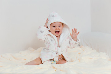 laughing baby girl in a Terry robe and diaper sitting on the bed after bathing. an eight-month-old Caucasian kid