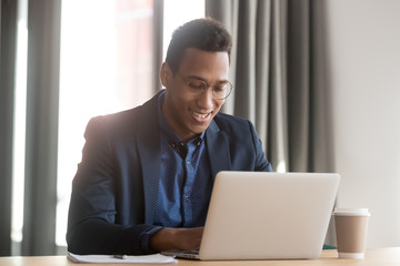 African ethnicity millennial businessman in glasses sitting in workplace smiling working on laptop,...