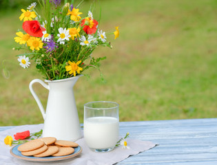 Wild flower bouquet in white metallic jug on vintage wooden table. Still life with bouquet of spring wildflowers, glass of milk, cookies. Spring, summer day. Healthy rustic breakfast. Copy space.