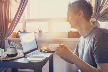 Young man working on laptop sitting near coffee table with cup of hot drink tea. Workplace near window on sutset or early morning. Working at home. Back light, soft selective focus. Copy space.