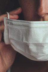 Detail photo of unrecognizable white woman putting on a white medical face mask to prevent the flu. Blurred background, focus on the mask. Coronavirus, COVID-19 quarantine. Lockdown concept