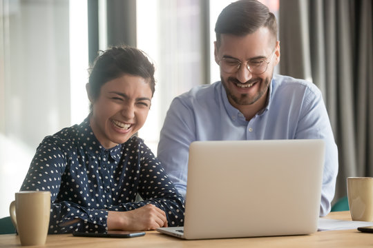 Multi-ethnic colleagues sit at desk working on project together use computer, telling jokes, laugh and humor brighten mood, key in creative thinking, increase effective workplace communication concept