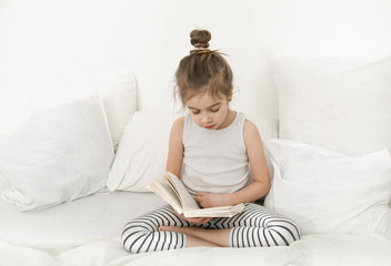 Cute little girl reading a book on the bed in the bedroom.