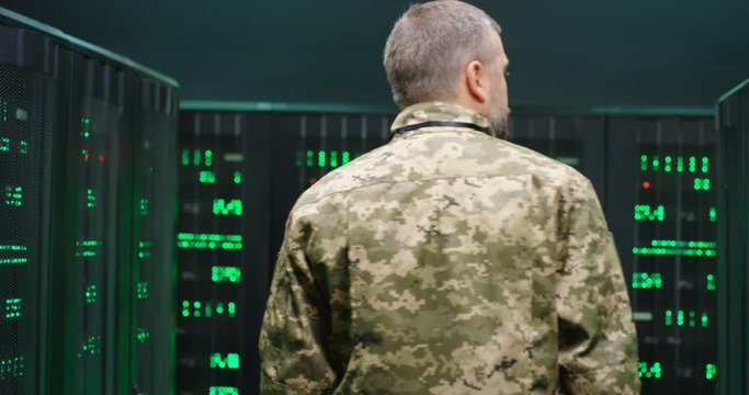 Rear of male Caucasian military worker walking the dark passage among servers with analytic secret data and green digits. Back view on man officer in camouflage in monitoring room.