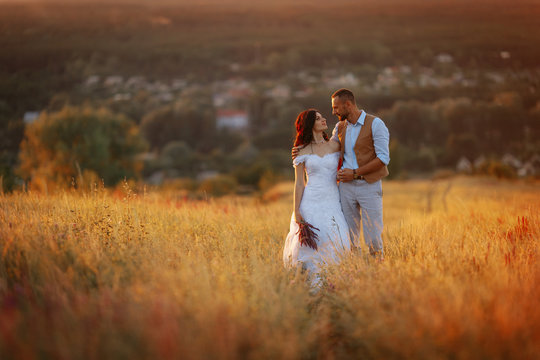 Young beautiful stylish couple in a flowering field of wheat on a road on a hill with a view of the city at sunset. Rustic wedding photo shoot. Bride and groom in a white dress with a train