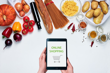 Female hands holding a tablet on white background next to groceries: bell peppers, dried chili, oregano, curry, allspice, onions, pasta, olive oil, baguette, tomatoes, pumpkin, potatoes