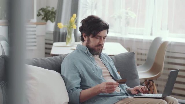 Tilt down shot of man sitting on couch in the living room, holding credit card and typing on laptop while buying goods in online store during quarantine at home