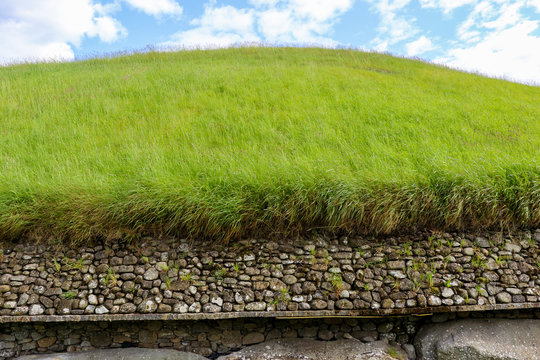 The lush green grass mound of Newgrange megalithic passage tomb in the Boyne Valley, Meath, Ireland contrasts with the bright blue sky and white clouds. The image is taken from the rear of the tomb