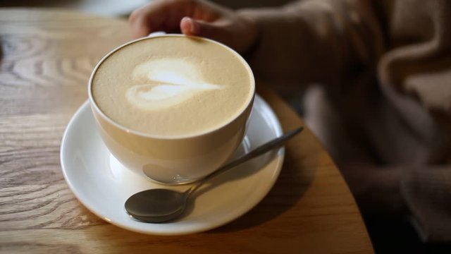 A Cup of coffee with a woman running her hand over it. The Cup contains a heart made of coffee foam.The Cup is on a saucer, and a teaspoon is on the saucer.Cafe