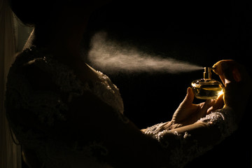 Mulher passando perfume no seu colo, com uma luz em meio a escuridão. Um frasco de perfume sendo segurado por uma pessoa - obrazy, fototapety, plakaty