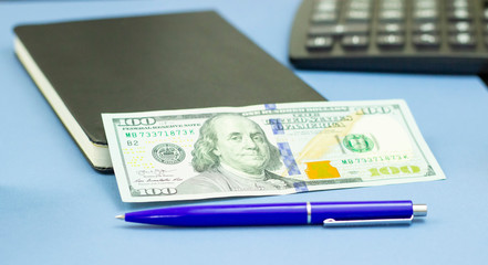 Notepad diary, US dollar bill and a ballpoint pen on a blue textured surface