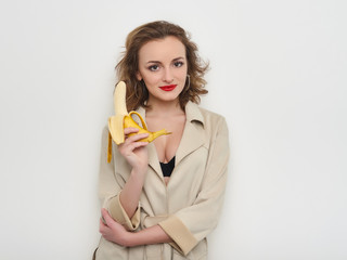 Sexy woman with red lips and curly hairdo in short coat and black bra playfully posing eating banana on white background
