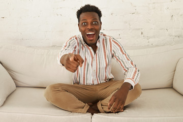 Portrait of emotional shocked young dark skinned guy relaxing at home, sitting comfortably on...