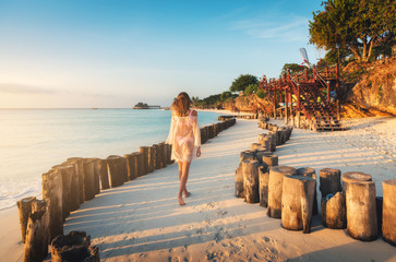 Beautiful young woman is walking on the sandy beach near blue sea at sunset. Summer travel....