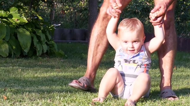 Grandfather teaches the toddler boy to walk. Baby boy making his first steps. The kid tries to walk barefoot on the grass in the summer in the back yard and holds dad's hands. Front view.