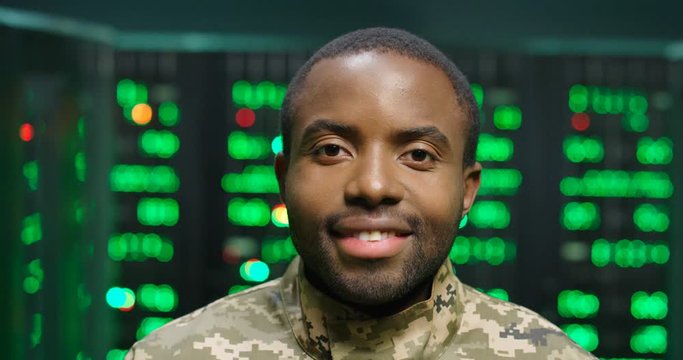Close up of African American military male officer in camouflage looking at camera in server room. Digital analytic center concept. Portrait of army official man smiling with servers on background.