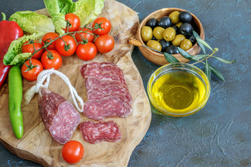 Fresh sliced fuet, tomatoes cherry and some vegetables on the kitchen cutting board. Olives in wooden bowl, olive oil in glass transparent bowl on the dark bacckground. Healthy food for snack.