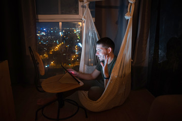Obraz na płótnie Canvas A man at home in a cozy hammock uses a tablet to communicate with family, friends and relatives via conference calls through messengers.