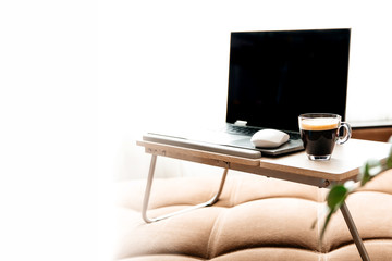 An open laptop and a Cup of coffee on a stand on the sofa.Empty white background on the left. home office. Working remotely. Work from home. Cozy office at home.