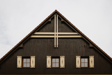 Fototapeta na wymiar Gable roof of cottage with cloudy sky in background.There are 3 glass windows with wooden decorated shutters. Gable is made of wooden planks with dominant wooden cross at its top.