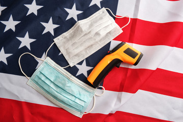 Used protective face masks and non-contact infrared thermometer on american flag background.