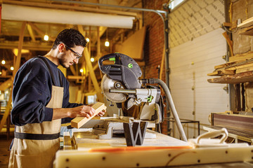 Fototapeta premium confident young carpenter work with mitre saw in factory, stand standing behind a workbench wearing uniform. industry, handicraft, carpentry concept