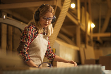 concentrated serious confident carpenter woman deals with piece of wood, she listen to music, protect her ears from noisy sounds in factory