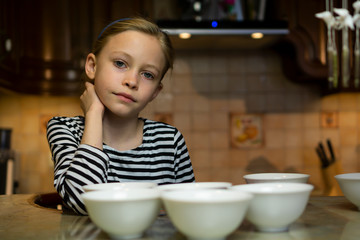 Cute caucasian blond child girl in a striped dress does not know what to cook sitting at cosy home kitchen table with many empty white plates bowls