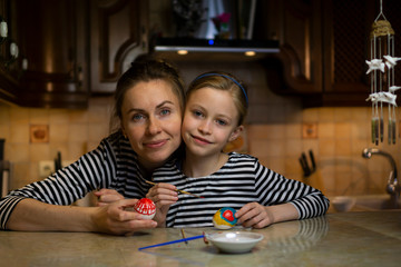 Beautiful caucasian mother and child daughter in striped dresses paint easter eggs sitting at table at cosy wooden kitchen at home. Family togetherness