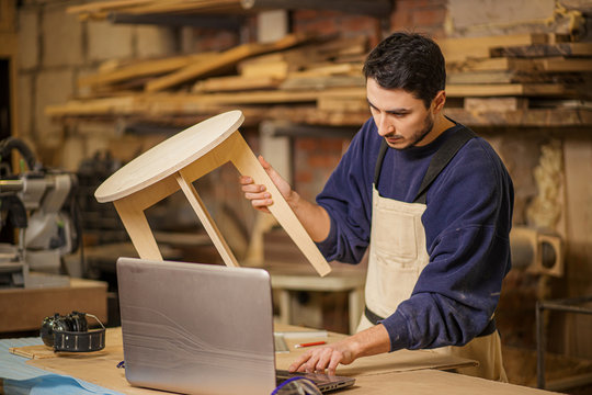 Young Furniture Maker Skillfully Make A Wooden Chair On A Workbench And Use Laptop While Working Alone In His Large Woodworking Shop, Handicraft And Carpentry Concept