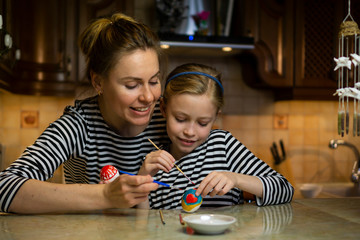 Caucasian mother helping child daughter in striped dress to hand paint easter eggs sitting at table at home cosy wooden kitchen. Family togetherness