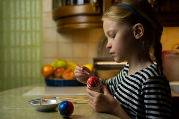 Beautiful blond child girl in striped dress hand painting easter egg with colorful paint at home kitchen table with fruit plate at background