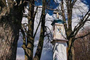 Przydrożna biała kapliczka uwieńczona krzyżem w otoczeniu drzew, a w tle błękit i obłoki
