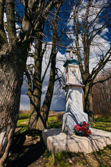 Przydrożna biała kapliczka uwieńczona krzyżem w otoczeniu drzew, a w tle błękit i obłoki © Marek Szymski