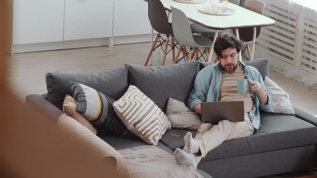 High angle shot of bearded Caucasian man sitting on comfortable couch at home, drinking tea from mug and browsing the Internet on laptop while doing online shopping