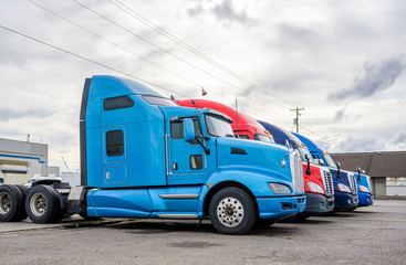 Different big rigs semi truck tractors standing on the warehouse parking lot waiting for next cargo loads for delivery