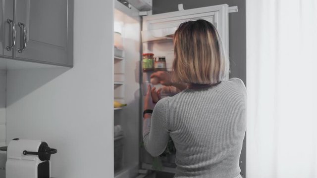 Woman opens refrigerator door in kitchen at home and takes two eggs and bottle of milk