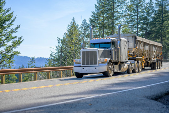 Gray classic big rig semi truck with vertical pipes transporting cargo in covered bulk semi trailer running on the mountain road with trees on the side