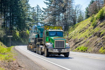 Green powerful big rig semi truck with oversize load yellow sign on the roof transporting crawler on the step down semi trailer running uphill on the winding mountain road