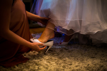 Mulher colocando o sapato branco de uma noiva no dia do seu casamento. Madrinha ajudando a noiva de vestido colocar o seu sapato de salto alto nos preparativos para realizar o sonho do matrimônio.  - obrazy, fototapety, plakaty