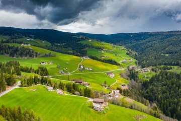 Fototapeta na wymiar Aerial view of improbable green meadows of Italian Alps, green slopes of the mountains, Bolzano, huge clouds over a valley, roof tops of houses, Dolomites on background, sunshines through clouds