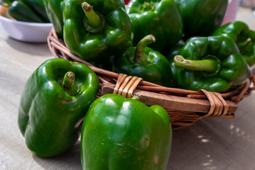 A small brown wicker basket with a bunch of green peppers in it and on a table next to the container. The skin on the bell pepper is thick and shiny.  The green vegetable has a long stalk on the top. 