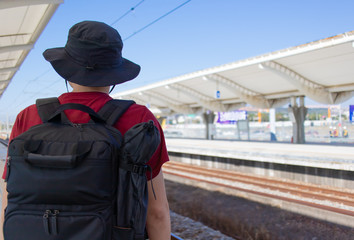Fototapeta premium 鉄道の駅のプラットフォームで電車を待つ黒いリュックサックを背負った男性の後ろ姿
