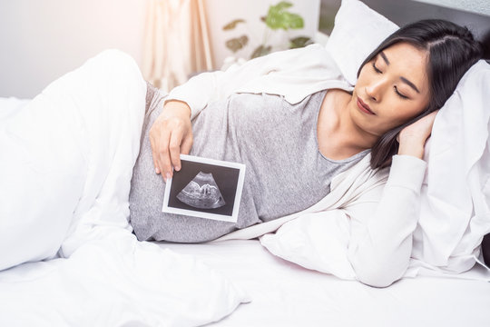 Beautiful asian pregnant woman laying on bed sideways, holding ultrasound baby picture on pregnant belly, resting and relaxing in bed room from hormone stress, comfy wearing stretch pants and cardigan