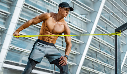Athletic man performs exercises using resistance band. Photo of strong man training in the city....
