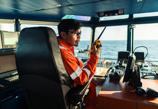 Filipino deck Officer on bridge of vessel or ship wearing coverall during navigaton watch at sea . He is speaking on portable GMDSS VHF radio, short range communication
