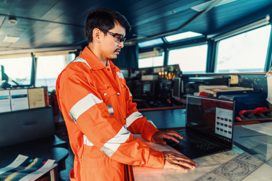 Filipino deck Officer on bridge of vessel or ship wearing coverall during navigaton watch at sea . He is using laptop, electronic paperwork at sea, concept of reporting